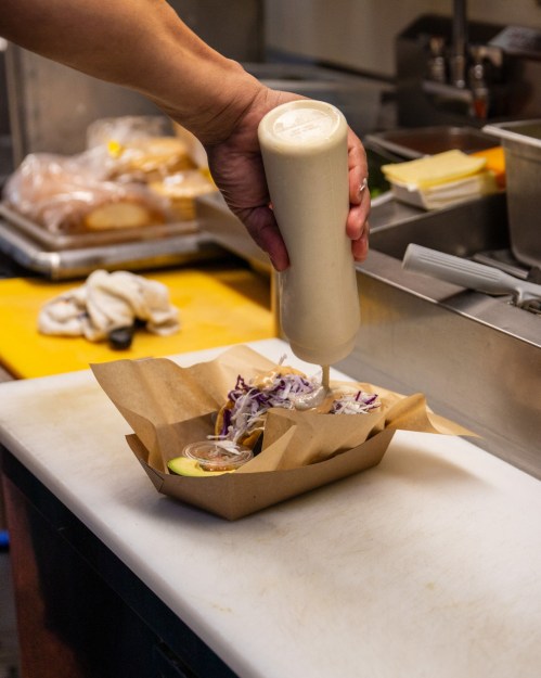 A hand holds a squeeze bottle, drizzling sauce over a tray of tacos with slaw and dipping sauce in a kitchen setting.