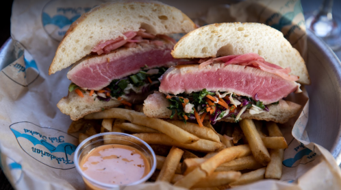 A sandwich with pink tuna, greens, and slaw, served with fries and a small cup of dipping sauce.