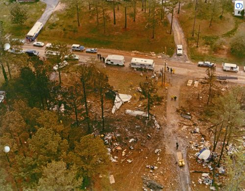 Aerial view of a disaster site with debris, emergency vehicles, and responders in a wooded area.