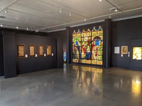 A gallery interior featuring stained glass windows and framed artworks on dark walls with polished concrete flooring.