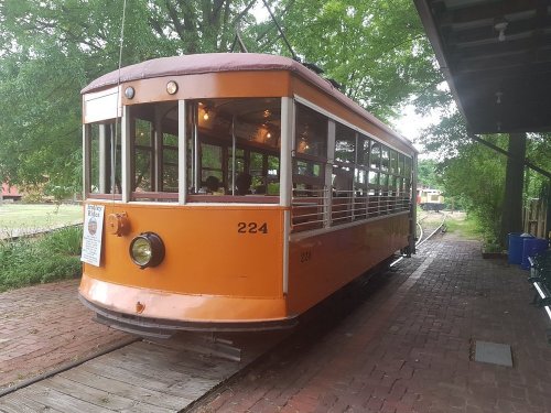 An orange vintage streetcar parked on a brick platform surrounded by greenery.