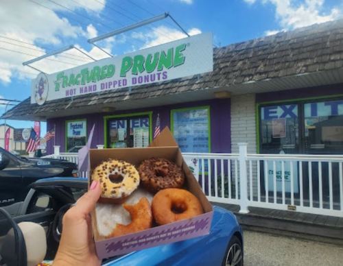 A hand holds a box of assorted donuts in front of a shop called "The Fractured Prune." Blue sky in the background.