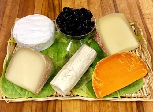 A wooden platter with various cheeses, including soft, hard, and orange varieties, alongside a bowl of dark fruit preserves.