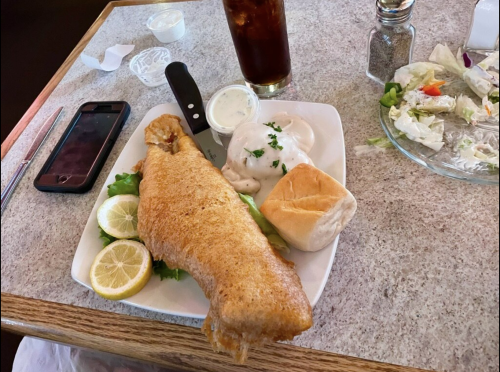 A plate with a large fried fish fillet, lemon slices, a roll, and a side of creamy sauce, with a salad and drink nearby.