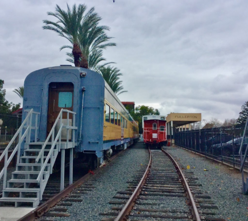 Two vintage train cars on tracks, with palm trees and a sign reading "Fullerton" in the background under a cloudy sky.