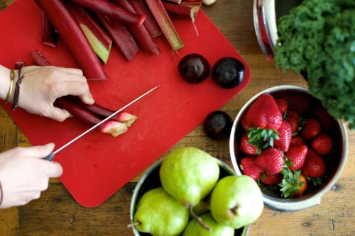 A person slices rhubarb on a red cutting board, surrounded by plums, strawberries, and green pears in bowls.