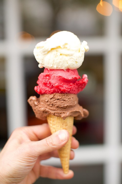 A hand holding a triple scoop ice cream cone with chocolate, strawberry, and vanilla flavors.