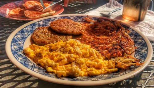 A plate of breakfast featuring scrambled eggs, hash browns, and sausage patties, with a fork in the background.