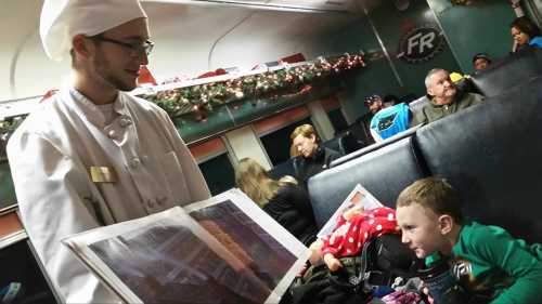 A chef in a white coat shows a menu to a young boy on a train, with festive decorations in the background.