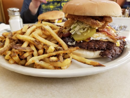A large burger stacked with fried chicken, lettuce, pickles, and bacon, served with a side of crispy French fries.
