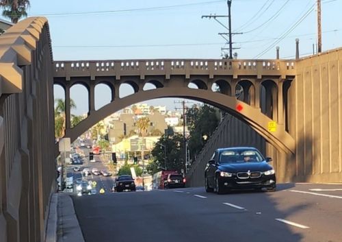 A black car drives down a street under a decorative bridge with arches, surrounded by buildings and power lines.