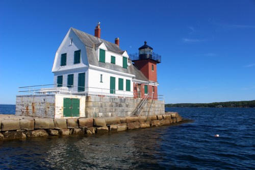 A white and red lighthouse on a rocky shore, surrounded by calm blue water under a clear blue sky.