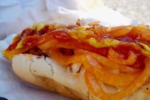 A close-up of a hot dog topped with onions, ketchup, and mustard, served in a soft bun.