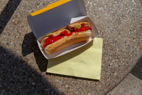A hot dog in a box topped with ketchup and mustard, resting on a yellow napkin on a textured surface.