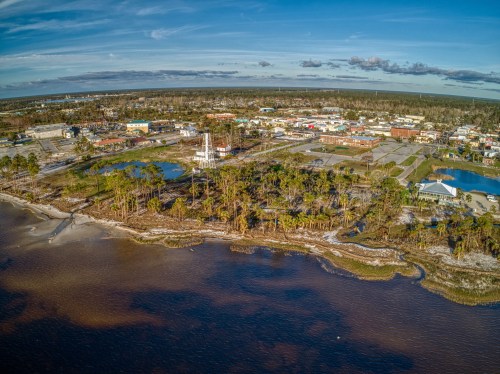Aerial view of a coastal town with water, trees, and buildings under a blue sky with scattered clouds.