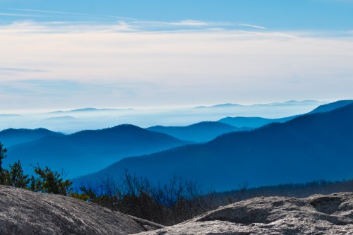 A serene landscape of layered blue mountains under a soft sky, with rocky foreground and distant peaks fading into the horizon.