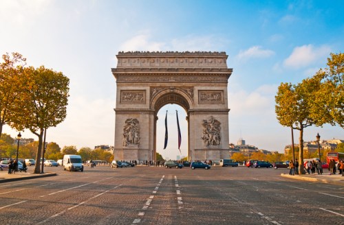 The Arc de Triomphe in Paris, framed by trees and a clear blue sky, with cars and people in the foreground.