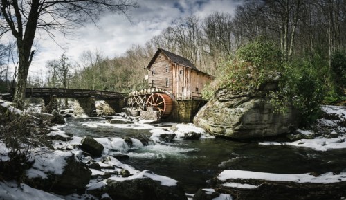 A rustic watermill beside a river, surrounded by trees and rocky terrain, with snow on the ground and a cloudy sky.