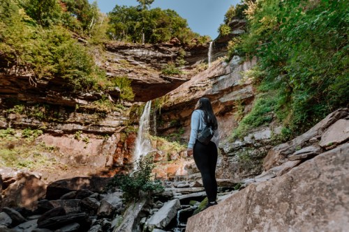 A person stands on rocks, gazing at a waterfall surrounded by lush greenery and cliffs.