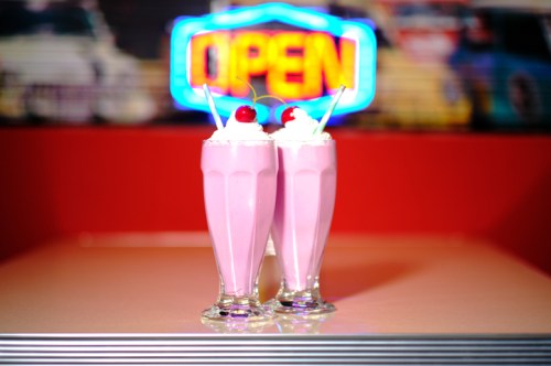 Two pink milkshakes topped with whipped cream and cherries, set against a vibrant "OPEN" neon sign.