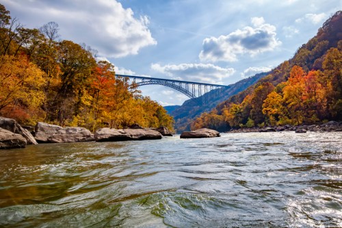 A scenic river view with autumn foliage and a bridge spanning the landscape under a partly cloudy sky.