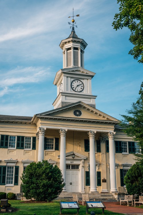 Historic building with a clock tower, white columns, and green shutters under a blue sky.
