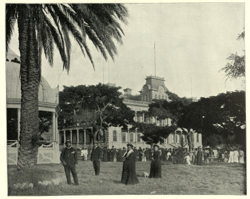 Historic black-and-white photo of a park with people, palm trees, and a large building in the background.