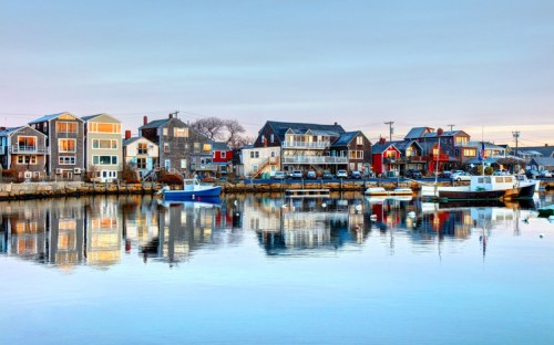 Colorful houses line a calm harbor, reflecting in the water under a clear sky. Boats are moored nearby.