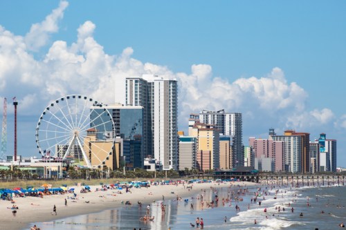 A beach scene with people, colorful umbrellas, and a skyline featuring tall buildings and a Ferris wheel under a blue sky.