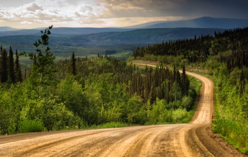A winding dirt road through lush green forests and rolling hills under a cloudy sky.