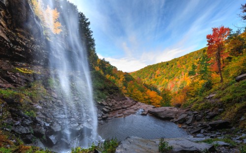 A stunning waterfall cascades into a serene pool, surrounded by vibrant autumn foliage and a clear blue sky.