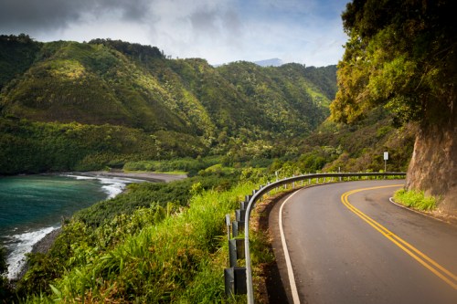 Winding road along a lush green coastline with mountains and a calm ocean in the background.