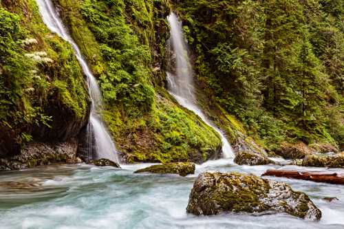 A serene landscape featuring two waterfalls cascading over moss-covered rocks into a flowing river surrounded by lush greenery.