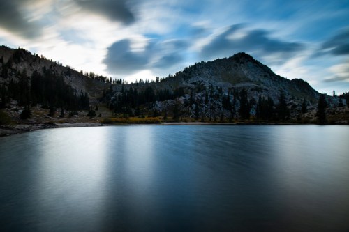 A serene lake reflects a cloudy sky, surrounded by mountains and trees in a tranquil landscape.