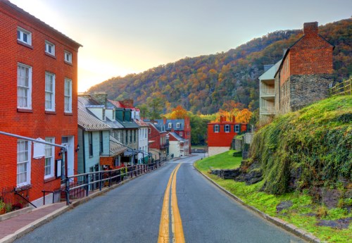 A scenic street lined with colorful buildings, leading towards a hillside with autumn foliage at sunset.