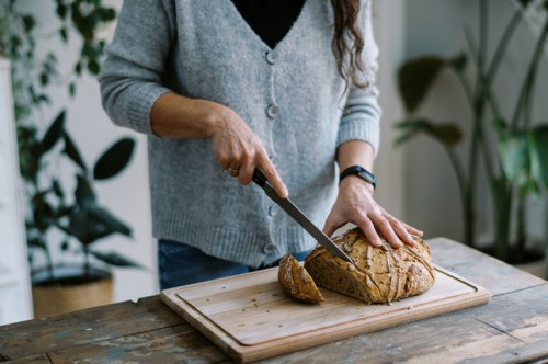 A person slices a loaf of bread on a wooden cutting board in a cozy, plant-filled kitchen.