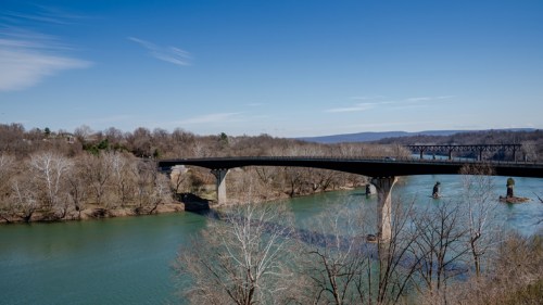 A bridge spans a river surrounded by trees, with a clear blue sky above and distant hills in the background.