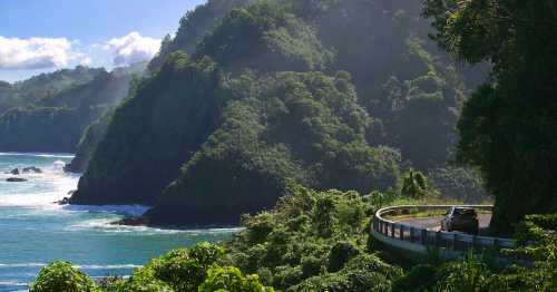 A scenic coastal road winds along lush green cliffs beside the ocean under a clear blue sky.