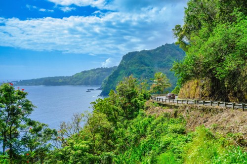 A scenic coastal road winds along lush green cliffs beside a calm ocean under a partly cloudy sky.