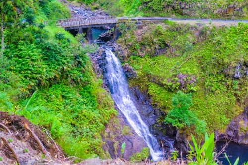 A scenic waterfall cascades down rocky terrain, surrounded by lush greenery and a bridge overhead.