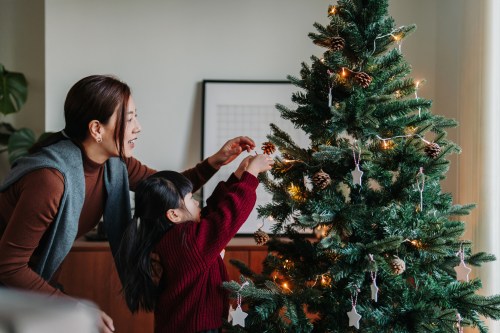 A woman and a child decorate a Christmas tree with ornaments and pinecones in a cozy living room.