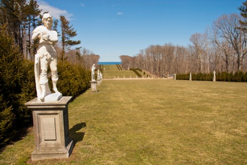 A landscaped garden with statues, green grass, and a view of the ocean in the distance under a clear blue sky.