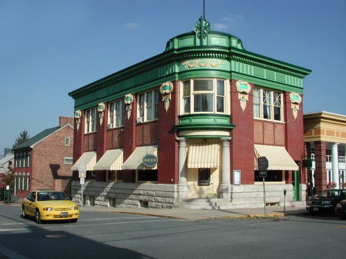 Historic brick building with green accents and decorative elements, featuring large windows and a yellow car passing by.