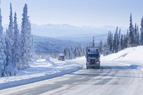 A truck drives on a snowy road surrounded by tall, snow-covered trees and mountains in the distance.