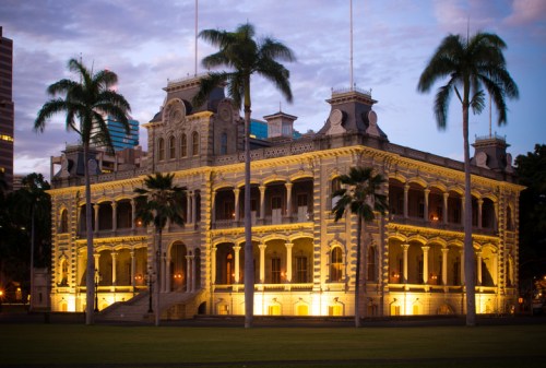 Historic building illuminated at dusk, surrounded by palm trees and city skyline in the background.