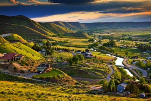A scenic view of rolling green hills, a winding river, and small houses under a dramatic sky at sunset.