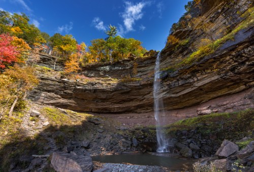 A scenic waterfall cascades down rocky cliffs, surrounded by vibrant autumn foliage and a clear blue sky.