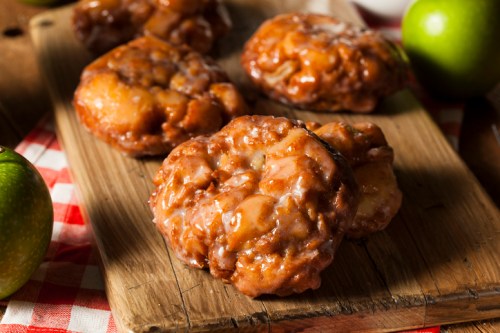 Freshly glazed apple fritters on a wooden board, surrounded by green apples and a checkered cloth.