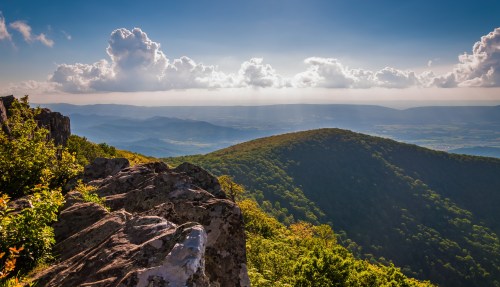 A scenic view of rolling green mountains under a blue sky with fluffy clouds, illuminated by sunlight.