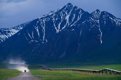 A rugged mountain landscape with snow-capped peaks, a dirt road, and a vehicle kicking up dust in the foreground.